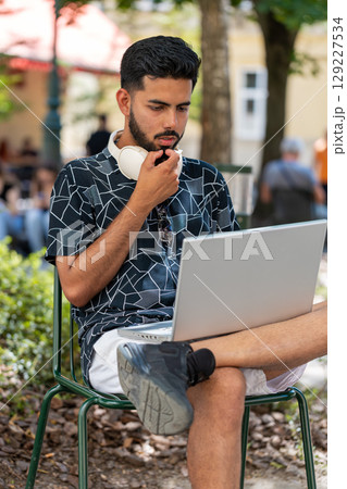 Indian man in headphones working online distant job with laptop browsing website chatting outdoors 129227534