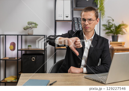 Upset young Caucasian businessman freelancer showing double thumbs down sitting at home office desk Upset young Caucasian businessman freelancer showing double thumbs down sitting at home office desk 129227546
