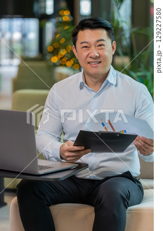 Chinese male entrepreneur in shirt with laptop analyzing graph documents reports in office lobby Chinese male entrepreneur in shirt with laptop analyzing graph documents reports in office lobby 129227580