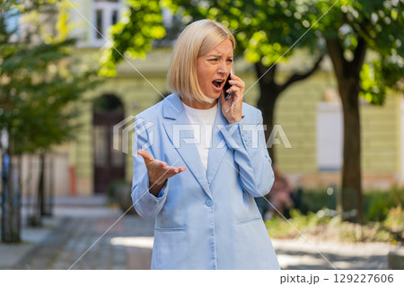 Angry mature Caucasian businesswoman arguing while talking on smartphone standing on city street Angry mature Caucasian businesswoman arguing while talking on smartphone standing on city street 129227606