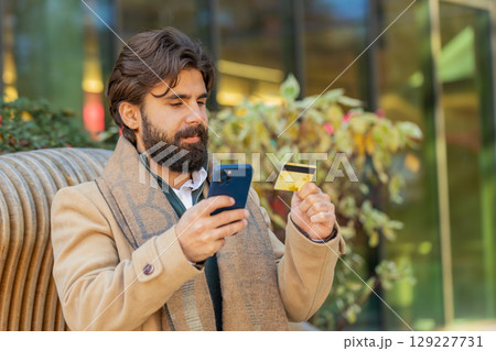 Indian man using credit bank plastic card and smartphone shopping online on downtown city street 129227731