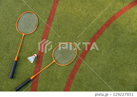 Two orange badminton rackets and a shuttlecock rest on a green sports field, captured from above. 129227851