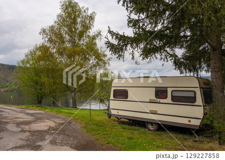 Vintage caravan parked near river under fir tree - peaceful spring camping scene in rural nature. 129227858
