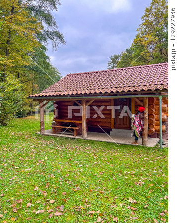 Woman in bright red cap and checkered blanket stands near wooden rustic cabin with cup in morning 129227936
