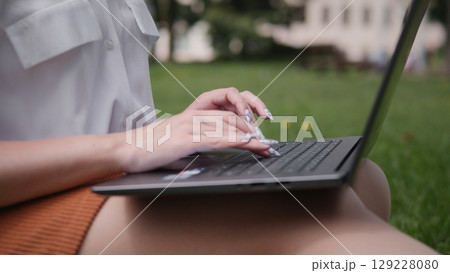 A woman engages in a productive meeting with friends outdoors, typing on a laptop in a green park. The bright summer day encourages collaboration and creativity in a natural setting. 129228080
