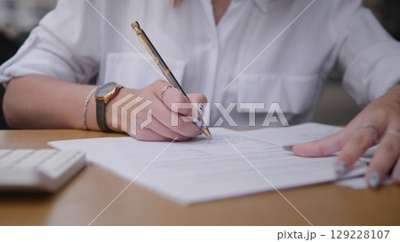 An individual diligently jotting down notes on paper with a pen at a cluttered desk full of papers and items 129228107