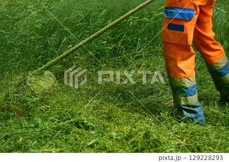 A park worker in an orange jumpsuit mows the grass with a motorized mower 129228293