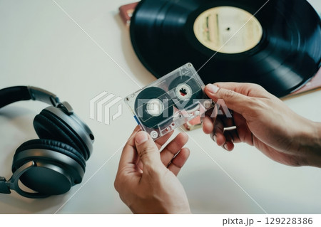 Hands hold a transparent cassette tape while a vinyl record and headphones lay nearby. The atmosphere evokes a sense of nostalgia and love for classic music styles. 129228386