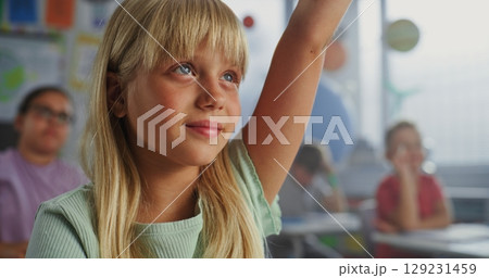 Close Up of Elementary School Girl Sitting at Desk, Raising Hand and Answering Teacher's Question 129231459