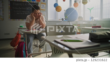 Exhausted Female Teacher Rubbing her Temples, Sitting Alone at Desk in Empty Classroom Exhausted Female Teacher Rubbing her Temples, Sitting Alone at Desk in Empty Classroom 129231622