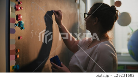 Female Teacher Writing Numbers and Multiplication Table on the Chalkboard, Teaching Primary School Students 129231692