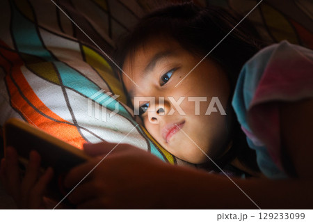 Asian girl using smartphone at night with glowing screen, concept of screen addiction and harmful blue light in children. 129233099