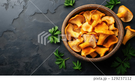 Fresh chanterelle mushrooms in wooden bowl with parsley leaves on dark stone background, rustic food concept. 129237112