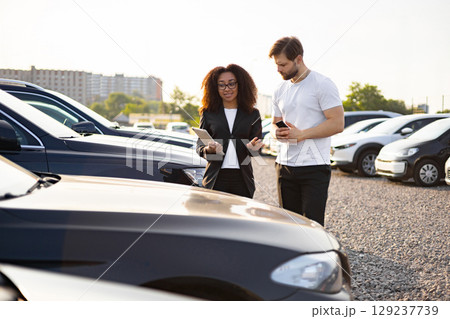 A female car dealer shows a car to a male customer in a car dealership parking lot during the daytime. A female car dealer shows a car to a male customer in a car dealership parking lot during the daytime. 129237739