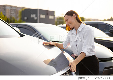 A smiling woman is looking at a new car, touching its hood, and considering a purchase at a dealership. 129237754