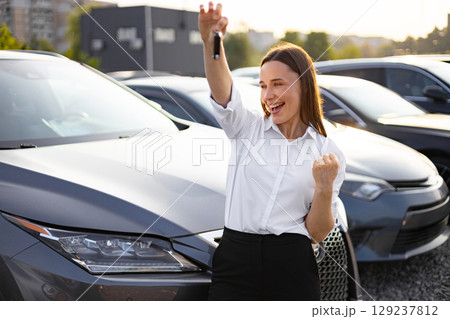 A joyful woman celebrates her car purchase, holding up the keys in excitement, standing near the new vehicle in a car dealership. A joyful woman celebrates her car purchase, holding up the keys in excitement, standing near the new vehicle in a car dealership. 129237812