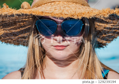 Close-up portrait of a young woman wearing a straw hat and blue sunglasses by the pool, enjoying a sunny summer day outdoors. Close-up portrait of a young woman wearing a straw hat and blue sunglasses by the pool, enjoying a sunny summer day outdoors. 129239194