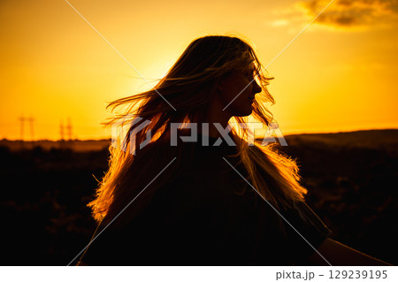 Silhouette of a young woman with long hair blowing in the wind, standing outdoors during sunset with a glowing orange sky in the background. Silhouette of a young woman with long hair blowing in the wind, standing outdoors during sunset with a glowing orange sky in the background. 129239195