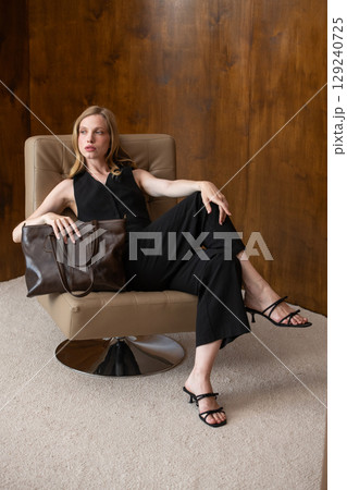 woman with wavy hair seated in a beige chair posing with a rich, dark brown leather tote bag. wearing sleeveless black top, trousers that drape gracefully to just above her black heeled sandals. 129240725