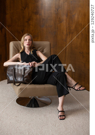 woman with wavy hair seated in a beige chair posing with a rich, dark brown leather tote bag. wearing sleeveless black top, trousers that drape gracefully to just above her black heeled sandals. woman with wavy hair seated in a beige chair posing with a rich, dark brown leather tote bag. wearing sleeveless black top, trousers that drape gracefully to just above her black heeled sandals. 129240727