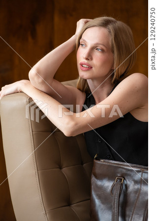 woman with wavy hair seated in a beige chair posing with a rich, dark brown leather tote bag. wearing sleeveless black top, trousers woman with wavy hair seated in a beige chair posing with a rich, dark brown leather tote bag. wearing sleeveless black top, trousers 129240750