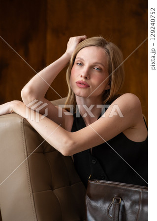 woman with wavy hair seated in a beige chair posing with a rich, dark brown leather tote bag. wearing sleeveless black top, trousers 129240752