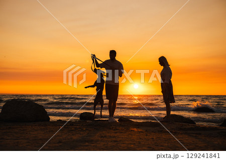 Man and woman with their German Boxer dog on the beach at sunset, playing as the dog jumps on its hind legs near the ocean waves. Man and woman with their German Boxer dog on the beach at sunset, playing as the dog jumps on its hind legs near the ocean waves. 129241841