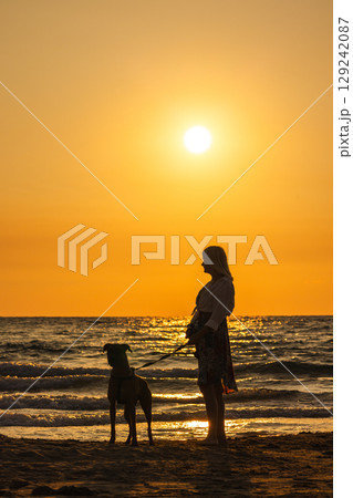 Woman standing with a dog on the beach at sunset, enjoying the evening by the sea shore Woman standing with a dog on the beach at sunset, enjoying the evening by the sea shore 129242087
