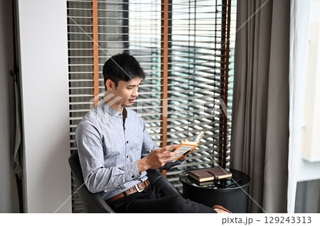 Adult businessman sitting by window reading a book in modern living space 129243313