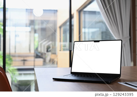 Laptop with blank screen on wooden desk in modern office interior with large glass windows 129243395