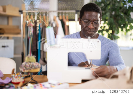 Working in sewing workshop - male tailor working at sewing machine making stitches on fabric 129243765