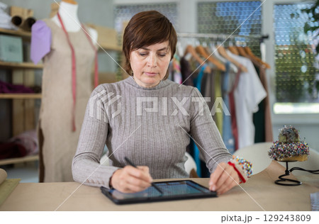 Female tailor draws sketch of future clothing on an electronic tablet. Working with modern gadgets in sewing workshop 129243809