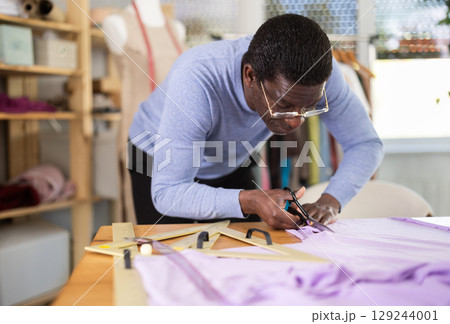 Male tailor cutting fabric with scissors while working in workshop 129244001