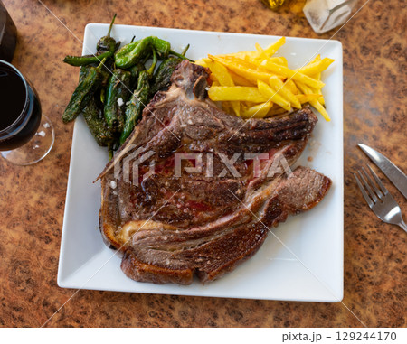 Grilled beef chop on bone, served with a vegetable side dish of fried potatoes and green padron peppers 129244170