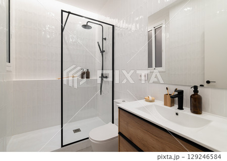 Modern bathroom with a glass shower, white tiles, wooden vanity, and minimalist decor. Clean and stylish design for contemporary living spaces. 129246584