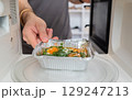 Person placing ready-to-eat meal in aluminum tray into a microwave oven for heating. 129247213