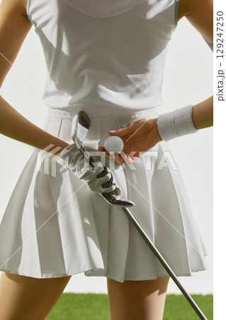 Woman in white golf outfit holding ball and iron club 129247250