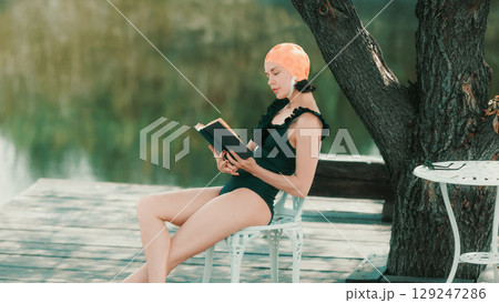 Woman in retro swimsuit reading book alone on chair near lake 129247286