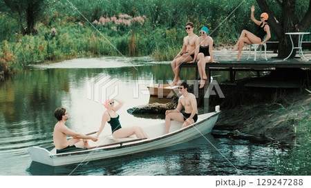 Group of friends in retro swimsuits relaxing on dock and in boat on calm river 129247288
