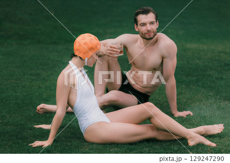 Woman in white swimsuit and man in black trunks sitting on lawn 129247290