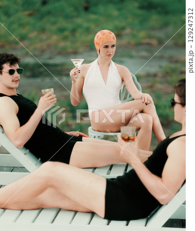 Woman in white swimsuit holding martini seated among two men in black vintage outfits 129247312