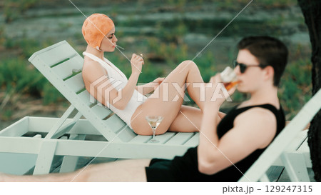 Woman in white swimsuit smoking by water with martini while man relaxes nearby Woman in white swimsuit smoking by water with martini while man relaxes nearby 129247315
