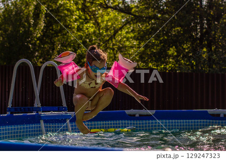 Child swimming in pool. A little girl in glasses and pink armbands jumps into a backyard pool on a sunny summer day, enjoying outdoor games and refreshing water treatments. 129247323