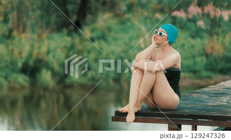 Woman in vintage swimsuit sitting alone on dock with peaceful expression Woman in vintage swimsuit sitting alone on dock with peaceful expression 129247326