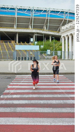 Two women jogging across red-striped pedestrian crossing near stadium 129247339