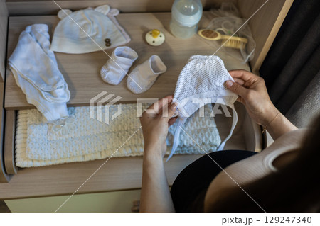 Pregnant woman holding baby hat while arranging clothes and accessories in drawer, preparing for childbirth and showing love and care for her unborn child. Preparing for motherhood. Pregnant woman holding baby hat while arranging clothes and accessories in drawer, preparing for childbirth and showing love and care for her unborn child. Preparing for motherhood. 129247340