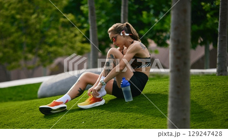Young woman putting shoes on sitting on grassy hill post-exercise in park 129247428