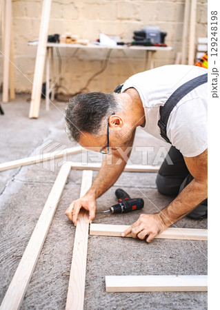 Mature male carpenter using electric screwdriver on a wooden plank outdoors Mature male carpenter using electric screwdriver on a wooden plank outdoors 129248198