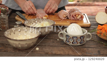 Turkey roll preparation process featuring a chef carefully stuffing turkey cuts with flavorful ingredients on a wooden countertop surrounded by fresh vegetables and seasonings 129248820
