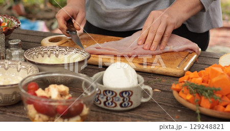 Expert chef prepares cut turkey fillet on wooden board, showcasing culinary skills in rustic kitchen, surrounded by fresh vegetables, herbs, and prepared ingredients for meal 129248821